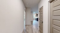 This single-floor unit presents a cohesive modern aesthetic defined by light wood-tone luxury vinyl plank flooring, crisp white walls, and black hardware accents carried consistently throughout the space. A central hallway connects the primary living areas, with five-panel interior doors adding architectural detail to the circulation path.

The kitchen serves as a functional centerpiece, offering two distinct configurations visible from the images: a galley-style prep corridor lined with dark walnut cabinetry, quartz countertops, and a full suite of stainless steel appliances including a refrigerator, range, microwave, and dishwasher. This opens into a more expansive kitchen area featuring a peninsula island with a matte black sink faucet and pendant lighting overhead. Multiple windows allow natural light to fill the space, offering views of surrounding greenery.

The living area is generously proportioned, with recessed lighting and a ceiling fan providing both illumination and air circulation. Bedrooms are well-sized with natural light provided by blinds-fitted windows. The bathroom features large-format marble-look tile surrounding the soaking tub, complemented by a wood-toned vanity and matte black fixtures, maintaining the unit's consistent contemporary character.