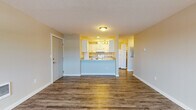 This single-floor unit at 210 South Farr Road in Spokane, Washington offers a clean, functional layout with consistent dark hardwood-style vinyl flooring throughout. The open living area is bright and spacious, featuring neutral-toned walls, a textured ceiling, a wall-mounted AC unit, and a large window that fills the room with natural light. A wall heater provides additional climate control, and a pet door is conveniently installed near the front entry.

The living space flows seamlessly into the kitchen area, which is separated by a half-wall peninsula with a light granite-style countertop. The kitchen is well-equipped with white shaker-style cabinetry, matching granite-look countertops, a tile backsplash, a deep double-basin sink with a brushed nickel faucet, a white electric range, microwave, and refrigerator. Track lighting illuminates the workspace, and ample cabinet storage lines both sides of the galley-style layout.

A secondary room features a sliding glass door that opens to a private outdoor area, allowing for natural light and easy outdoor access. The bathroom is neatly finished with a white vanity with granite-style countertop, a combination tub and shower, and coordinating hardwood-style flooring, completing this thoughtfully updated unit.
