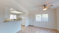 This single-floor space offers a clean, open-concept layout that seamlessly connects the living area and kitchen. The living area features light hardwood-style flooring, crisp white walls, and a ceiling fan with a light fixture, contributing to a bright and airy atmosphere. A large window allows natural light to fill the room while offering views of surrounding greenery, enhancing the overall sense of openness. A white paneled door provides access to the exterior.

The kitchen is accessible via an open archway and is defined by white cabinetry, a raised breakfast bar counter with a warm wood-toned surface, and a white refrigerator. A skylight panel above the kitchen workspace provides additional natural illumination, keeping the cooking area well-lit throughout the day. The consistent white color palette across both spaces creates a cohesive and neutral aesthetic throughout.

Overall, the space presents a functional and well-maintained interior that feels open and inviting, with practical features and a simple design that maximizes natural light and flow between the living and kitchen areas.