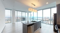 This high-rise unit is defined by its striking floor-to-ceiling glass windows, which wrap around the main living and bedroom areas to deliver expansive, panoramic city views. Light wood-look flooring runs throughout the open-plan living and kitchen spaces, enhancing the bright, airy atmosphere created by the abundant natural light.

The kitchen is a standout feature, anchored by a large center island with a dark quartz countertop, an undermount sink, and a rectangular pendant light overhead. Cabinetry in a warm gray-wood finish extends from floor to ceiling, complemented by a white subway tile backsplash and a full suite of stainless steel appliances, including a refrigerator, range, and microwave.

The living area is generously proportioned, with light hardwood flooring and unobstructed views of the urban skyline. The bedroom features plush carpeting and direct access to the floor-to-ceiling windows, continuing the theme of elevated city views.

Two bathrooms serve the unit. One includes a tub-shower combination surrounded by classic white subway tile and brushed nickel fixtures. The other features a walk-in tiled shower, a floating dark-wood vanity with an integrated sink, and wood-look flooring, accessible via a sliding barn-style door adjacent to a walk-in closet.