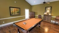 Located along the I-20 Frontage Road in Vicksburg, Mississippi, this single-story space offers a welcoming and well-appointed interior designed with both comfort and entertainment in mind. The standout feature is a spacious game room showcasing soaring vaulted ceilings that lend an open, airy feel to the space. Rich hardwood flooring runs throughout, adding warmth and a sense of classic elegance to the room. A full-size pool table with an orange felt surface serves as the centerpiece, making this an ideal gathering spot for recreation and leisure. The walls are finished in a warm olive green tone, complemented by crisp white chair rail molding that adds a refined architectural detail. Natural light filters in through a window fitted with blinds, brightening the space while maintaining privacy. A ceiling fan provides comfortable airflow overhead. The room is further enhanced by tasteful decorative touches, including framed artwork depicting an autumnal landscape, an ornate wooden dresser, and a carved accent cabinet, all contributing to a cozy yet sophisticated ambiance. This space is well-suited for entertaining guests or enjoying casual downtime in a characterful, inviting environment.