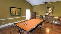 Located along the I-20 Frontage Road in Vicksburg, Mississippi, this single-story space offers a welcoming and well-appointed interior designed with both comfort and entertainment in mind. The standout feature is a spacious game room showcasing soaring vaulted ceilings that lend an open, airy feel to the space. Rich hardwood flooring runs throughout, adding warmth and a sense of classic elegance to the room. A full-size pool table with an orange felt surface serves as the centerpiece, making this an ideal gathering spot for recreation and leisure. The walls are finished in a warm olive green tone, complemented by crisp white chair rail molding that adds a refined architectural detail. Natural light filters in through a window fitted with blinds, brightening the space while maintaining privacy. A ceiling fan provides comfortable airflow overhead. The room is further enhanced by tasteful decorative touches, including framed artwork depicting an autumnal landscape, an ornate wooden dresser, and a carved accent cabinet, all contributing to a cozy yet sophisticated ambiance. This space is well-suited for entertaining guests or enjoying casual downtime in a characterful, inviting environment.