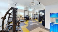 Located at 98 N 1st St in San Jose, CA, this fitness center offers a thoughtfully designed exercise space built to support a wide range of workout routines. The room features soaring ceilings that create an open, airy atmosphere, complemented by large windows along the upper walls that flood the space with abundant natural light and offer views of the surrounding greenery. Ceiling fans and modern circular pendant lighting are suspended overhead, ensuring both comfort and visibility throughout.

The flooring features a dynamic patchwork of yellow, gray, and neutral carpet tiles that add a vibrant, energetic character to the space. Full-length wall mirrors strategically expand the sense of space and allow users to monitor their form during workouts.

The facility is comprehensively equipped for both strength and cardio training. Cardio offerings include treadmills and additional aerobic machines, while the strength zone features a multi-functional cable machine, a power rack with a bench, and a dedicated TRX functional training rig complete with a punching bag, medicine balls, kettlebells, and resistance bands. A dumbbell rack rounds out the free weights section. The space also includes modern specialty equipment, making it a versatile and fully capable fitness destination.