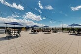 Located at 60 S Main St in Layton, Utah, this rooftop terrace offers an expansive open-air experience designed for relaxation and social gatherings. The space features a generously sized, tiled deck that accommodates multiple seating arrangements, each shaded by navy blue market umbrellas and furnished with durable cast-iron bistro tables and chairs. Overhead, strands of café-style string lights crisscross the terrace, creating a warm and inviting atmosphere for evening use.

A built-in outdoor grill station with tiled surround serves as a focal point for outdoor cooking and entertaining. Adjacent to the seating area, a raised hot tub provides a central amenity, surrounded by a structured deck platform with stair access. Raised planter beds are positioned along the perimeter, adding greenery and architectural definition to the space.

The terrace is enclosed by a perimeter railing system that ensures safety while preserving the stunning panoramic views. Sweeping vistas of the Wasatch Mountains frame the horizon, with the surrounding Layton cityscape visible in all directions. The open sky and abundant natural sunlight enhance the overall ambiance, making this rooftop terrace a standout outdoor destination in the heart of downtown Layton.
