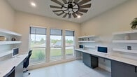 This contemporary business center is thoughtfully designed to support focused, productive work in a clean and welcoming environment. The space is anchored by a long, built-in workstation finished in white and charcoal gray, accommodating multiple users simultaneously with individual computer monitors and ample desk surface. Floating white shelves mounted above the workstations provide convenient display and storage space, accented with decorative objects and greenery that add a touch of warmth to the otherwise minimalist aesthetic.

Large windows fitted with adjustable roller shades line one wall, flooding the space with natural light while offering views of lush outdoor greenery and landscaped surroundings. The light-toned, large-format tile flooring enhances the bright, open atmosphere throughout. A striking windmill-style ceiling fan serves as a distinctive architectural focal point overhead, adding a modern-rustic character to the otherwise sleek interior.

A glass-paneled interior door connects the business center to an adjacent communal area, maintaining visual openness while providing acoustic separation. The neutral warm-white walls and recessed lighting contribute to a calm, professional ambiance. This well-appointed business center offers a functional and stylish setting for residents or guests seeking a dedicated workspace.