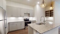 This single-floor residence presents a thoughtfully designed open-concept layout that seamlessly connects the kitchen and living areas. The kitchen is a standout feature, centered around a generous island with granite countertops and an undermount sink paired with a pull-down faucet. Tall gray shaker-style cabinetry lines the walls, complemented by a white tile backsplash and stainless steel appliances including a refrigerator, electric range, and dishwasher. Geometric pendant lights suspended above the island add a modern, decorative touch. Open wood shelving provides additional display and storage space near the entry.

Wood-look plank flooring flows consistently throughout the living areas and bedrooms, contributing to a cohesive and contemporary aesthetic. The living area receives natural light through large windows and features a ceiling fan. Bedrooms are bright and well-proportioned, with windows fitted with blinds. The walk-in closet offers multi-level shelving and hanging rods for organized storage.

The bathroom is finished with marble-look tile flooring, a dual-sink vanity topped with granite, framed mirrors, and gray cabinetry, delivering a clean and polished look throughout.