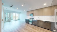 This well-appointed two-floor residence offers a thoughtfully designed layout spread across a compact yet efficiently utilized footprint. The lower level houses an open-plan kitchen and living area, while the upper level accommodates the bedroom, bathroom, and closet.

The kitchen is a standout feature, outfitted with high-gloss dark cabinetry contrasted by warm taupe upper cabinets and a marble-style tile backsplash. Stainless steel appliances, including a gas range with microwave overhead and a full-size French door refrigerator, complete the contemporary look. Light hardwood flooring flows throughout, enhancing the clean, modern aesthetic.

The living area benefits from expansive floor-to-ceiling sliding glass doors that open onto a glass-railed balcony, offering sweeping views of the surrounding cityscape and parkland beyond. Abundant natural light fills the space throughout the day.

The bedroom features its own balcony access via a glass sliding door, framing urban views and continuing the bright, airy feel of the home. The bathroom is finished in a clean, minimalist style with a floating wood-toned vanity, backlit mirror, and a glass-enclosed walk-in shower, completing the modern design language present throughout this residence.