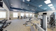 The fitness center at Capital Place At Southwood is a thoughtfully designed, single-floor workout facility that combines functionality with a welcoming atmosphere. The space features a striking deep blue ceiling accented by recessed lighting and ceiling fans, creating a dynamic contrast against the light-toned walls and neutral-patterned carpet flooring below. Natural light filters in through a row of windows along one wall, brightening the interior and providing an energizing environment for exercise. The cardio section is well-appointed with treadmills, elliptical machines, and stationary bikes, all positioned to take advantage of the natural light. Strength training is also well represented, with adjustable benches, free weights, resistance equipment, and stability balls available throughout the space. A wall-mounted television adds a convenient entertainment option during workouts. The layout is open and spacious, allowing for comfortable movement between equipment zones. Ceiling fans maintain airflow throughout the facility, contributing to a comfortable workout experience. The overall ambiance is clean, modern, and purpose-built, making the fitness center a practical and inviting amenity for residents of Capital Place At Southwood.