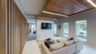 Located at 6101 N Keystone Ave in Indianapolis, Indiana, this thoughtfully designed single-floor community space blends modern aesthetics with functional versatility. The interior is anchored by a spacious lounge area finished with large-format tile flooring, a striking wood-slat ceiling with integrated linear lighting, and an array of oversized windows that flood the space with natural light and offer views of the surrounding streetscape. Vertical wood slat partitions elegantly define zones while maintaining an open, airy feel throughout.

A dedicated seating lounge features plush sectional sofas, mid-century modern accent chairs, and glass-topped coffee tables arranged around a wall-mounted television — ideal for casual gatherings or relaxation. Adjacent to this is a formal conference area outfitted with a solid wood dining table, upholstered navy chairs, gold pendant lighting, and its own wall-mounted display screen, all set beneath a warm wood ceiling detail.

The café-style nook offers a charming retreat with bold yellow banquette seating, marble-top bistro tables, decorative pendant lights, and a geometric accent wall. A built-in beverage station with light wood cabinetry, a beverage cooler, and coffee service adds practical convenience. The overall ambiance is clean, contemporary, and welcoming — a cohesive blend of warm wood tones, neutral walls, and curated furnishings.