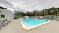 This single-floor property's most prominent feature is its expansive outdoor swimming pool, which serves as the centerpiece of the exterior space. The pool is well-maintained, showcasing clear, blue-tinted water and a smooth surrounding deck that offers ample room for lounging and recreational activity. A white metal safety fence encloses the pool area, providing both security and a clean, classic aesthetic. Life-saving equipment is mounted along the fence, indicating the space is thoughtfully equipped for safety. The pool area is framed by a rich backdrop of mature tropical vegetation, including towering palm trees and broad-canopied oak trees, which provide natural shade and privacy while lending the space a lush, subtropical character. The surrounding landscaping creates a serene and resort-like ambiance. The interior areas of the property were not well-lit in the available imagery, limiting detailed observation; however, the overall setting strongly suggests a community or residential property situated in a warm, tropical climate, likely in Florida, designed with outdoor living and leisure as a central focus.
