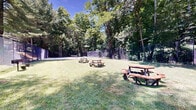 Located at 340 Media Station Rd in Media, Pennsylvania, this outdoor recreational area combines a generous picnic lawn with dedicated pickleball courts, creating a well-rounded amenity space set against a lush, natural backdrop. The grounds are framed by tall, mature trees that provide ample shade and a sense of seclusion, while the open, well-kept grassy lawn offers plenty of room for casual gatherings and outdoor activities. Multiple wooden picnic tables are spread throughout the lawn area, providing comfortable spots for dining and socializing. A charcoal grill is also available, making the space well-suited for outdoor cookouts. The pickleball courts are a standout feature, finished in a blue and green hard-court surface and enclosed by chain-link fencing for safe, uninterrupted play. The courts are equipped with a regulation net and accommodate both singles and doubles play. A small utility structure is visible near the perimeter, and the surrounding residential building provides convenient access to the amenity area. The overall ambiance is relaxed and inviting, with abundant natural light filtering through the tree canopy and a peaceful, park-like atmosphere that encourages both active recreation and leisurely outdoor enjoyment throughout the warmer months.