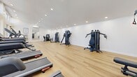 Located in Fullerton, CA, this fitness center offers a well-appointed exercise space designed for a comprehensive workout experience. The room features soaring two-story ceilings lined with a continuous row of clerestory windows along one wall, flooding the interior with abundant natural light and providing views of the surrounding outdoor pergola and mature trees. Light-toned hardwood flooring runs throughout, lending a clean and modern aesthetic to the space. Recessed lighting supplements the natural light, ensuring the room remains bright and inviting at all hours. The cardio zone is well-stocked with multiple treadmills, an elliptical trainer, a stationary bike, a stair climber, and a rowing machine, thoughtfully arranged to maximize open floor space. Along the opposite wall, a lineup of strength training cable and weight machines provides a full range of resistance exercises. A large wall-mounted mirror enhances the sense of space and supports form-checking during workouts. A wall-mounted split air conditioning unit ensures climate comfort year-round, while a water fountain offers convenient hydration. The layout is open and uncluttered, allowing for ease of movement between equipment and making this fitness center a functional and welcoming space for residents.