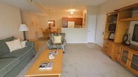 This single-floor residence showcases an open-concept floor plan that seamlessly connects the living, dining, and kitchen areas into one cohesive, functional space. The living area is anchored by plush wall-to-wall carpeting in a warm neutral tone, complemented by a wooden coffee table and a patterned accent chair with a coordinating throw pillow, creating a comfortable and inviting atmosphere.

The dining area is positioned adjacent to the living space and features a wooden table with matching chairs, illuminated by a classic chandelier overhead. A floral centerpiece and framed artwork on the walls add decorative touches that enhance the welcoming ambiance.

The kitchen is partially open to the main living area, separated by a raised countertop that serves as a natural divider while maintaining visual connectivity. Warm wood-toned cabinetry lines the kitchen walls, and a white refrigerator is visible, contributing to a clean, functional layout. A ceiling-mounted light fixture provides additional illumination.

Near the kitchen, a white entry door with a tall wooden storage unit alongside it provides both a practical entryway and added storage. The overall design emphasizes an open, airy feel with consistent warm tones throughout.