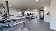 Located at 5600 Orangethorpe Ave in La Palma, California, this fitness center offers a well-appointed exercise space designed for efficient, focused workouts. The room features a clean, modern aesthetic with light gray walls, white trim, and durable dark concrete-style flooring that provides both a sleek look and practical durability underfoot.

The space is equipped with a comprehensive selection of cardio machines, including multiple treadmills, an elliptical trainer, a rowing machine, and a stationary bike, providing users with a variety of workout options. A large wall-mounted mirror runs along one side of the room, visually expanding the space and allowing users to monitor their form during exercise.

Natural light filters in through multiple windows, including a set of wide windows and a glass-paneled door, creating a bright and energizing atmosphere. A wall-mounted mini-split air conditioning unit ensures comfortable climate control year-round. Recessed ceiling lighting supplements the natural light, keeping the space well-illuminated at any time of day. A wall-mounted sanitizing station is also present, supporting cleanliness and hygiene. The black-framed glass entry door adds a modern, polished touch to the overall design, making this fitness center a functional and inviting facility.