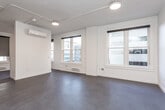 This single-floor unit offers a clean, modern aesthetic defined by an open-concept layout and a crisp white palette throughout. The expansive main living area features smooth concrete-style flooring, exposed conduit along the ceiling with track lighting, and a wall-mounted split HVAC unit, contributing to an industrial-modern character. Multiple double-hung windows line the exterior walls, flooding the space with natural light and offering views of the surrounding urban streetscape. Baseboard heating runs beneath the windows, providing an additional source of warmth. The kitchen is thoughtfully positioned within the open floor plan and is outfitted with white shaker cabinetry, granite-style countertops, and a full suite of stainless steel appliances including a refrigerator, range, microwave, and dishwasher. A stacked washer and dryer unit is also present, adding in-unit laundry convenience. The bathroom features a glass-enclosed shower with textured privacy panels. A dedicated bedroom area, separated from the main living space, benefits from its own window exposure, providing natural light and ventilation. The overall design balances functionality with a contemporary urban sensibility, making it a practical and well-appointed living space.