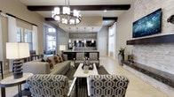 This sophisticated single-floor space blends contemporary design with refined architectural details to create an inviting and visually striking environment. The main lounge area is anchored by a statement chandelier with cylindrical shades, complemented by dark exposed ceiling beams that add depth and contrast to the light, neutral palette of the walls and flooring. Large windows allow natural light to fill the space, enhancing its open and airy feel.

The seating area is thoughtfully arranged with a plush sofa adorned with bold accent pillows, a sleek white rectangular coffee table, and a collection of patterned armchairs featuring a striking geometric hexagonal print. A dark area rug grounds the seating arrangement, adding visual warmth and definition.

Toward the rear of the space, a well-appointed wet bar or kitchenette is outfitted with dark cabinetry, a mosaic tile backsplash, and pendant lighting hung above the counter, creating an intimate and functional entertaining zone. To one side, a wall-mounted flat-screen television sits above a stone-clad surface, adding a modern media focal point. Lush tropical plants are positioned throughout, introducing natural elements that soften the space and enhance its upscale ambiance.