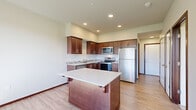 This one-bedroom, one-bathroom unit at Taylor Place in Stanwood, Washington offers a thoughtfully designed single-floor layout that maximizes its compact footprint. The kitchen serves as a central highlight, showcasing rich dark-wood cabinetry with sleek bar-pull hardware, light-toned countertops, and a mosaic tile backsplash. A full suite of stainless steel appliances—including a refrigerator, electric range, built-in microwave, and dishwasher—adds a modern, polished look. A freestanding kitchen island with a white countertop provides additional workspace and casual seating. Light hardwood-style flooring and recessed lighting throughout the kitchen and main living areas contribute to a bright, open atmosphere. The bedroom offers comfortable carpeting, a closet with built-in shelving, and a window that brings in natural light and outdoor views of the surrounding landscape. A hallway connects the spaces efficiently, leading to the full bathroom, which features wood-toned cabinetry, a combined tub and shower, and matching hardwood-style flooring. The overall aesthetic is clean and contemporary, with consistent warm-toned finishes creating a cohesive feel throughout.