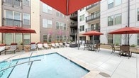 Located at 1005 W Main St in the heart of Durham, NC, this outdoor amenity deck is a thoughtfully designed communal space that combines modern urban architecture with resort-style leisure. The centerpiece is a generously sized swimming pool with crystal-clear blue water, accessible via stainless steel entry rails and surrounded by wide, large-format tiled pool decking that provides ample room for relaxation and movement.

Rows of cushioned chaise lounges fitted with bold red headrests are arranged poolside, many shaded by signature red market umbrellas. Multiple pergola structures offer additional shaded seating areas, outfitted with comfortable lounge furniture ideal for gathering. A fully equipped outdoor kitchen station features a large stainless steel grill, perfect for alfresco dining.

A dedicated turf lawn area provides a lush green retreat strung with bistro lights overhead, bordered by raised planters with seasonal flowering trees and ornamental shrubs that add natural color and texture. Dining tables with umbrella shade are positioned across the deck, creating distinct zones for lounging, dining, and socializing. The multi-story building façade, clad in a mix of brick, panel siding, and large windows with private balconies, frames the courtyard and contributes to the space's vibrant, contemporary atmosphere.
