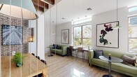 Located at 135 State Street in West Columbia, SC, this thoughtfully designed coffee bar offers a welcoming and functional layout across a single floor. The space is immediately striking thanks to its soaring ceilings adorned with richly toned exposed wood beams, which contrast beautifully against crisp white walls and light wood-look flooring throughout.

The lounge area features comfortable sage green sofas, sleek bistro-style tables, and an assortment of seating arrangements suited for casual gatherings or individual work. Large windows flood the space with natural light, framing views of the surrounding greenery and creating a warm, airy ambiance. Pendant lighting in muted tones adds a contemporary touch and layers the illumination throughout the room.

Curated large-format botanical artwork adorns the walls, contributing to the space's refined yet approachable aesthetic. A dining area with a natural wood table and bar-height seating offers additional flexibility for guests.

The coffee bar itself is a standout feature, anchored by a herringbone-tiled accent wall, stainless steel appliances, clean white countertops, and open wood shelving styled with plants and artisan goods. Built-in shelving in a soft sage tone provides display and storage. The space is entered through elegant white-framed glass French doors, setting an inviting tone from the moment of arrival.