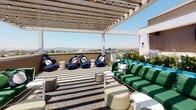 This rooftop terrace is a well-appointed outdoor amenity space designed for both relaxation and entertainment. The layout is anchored by a slatted pergola strung with globe lights, providing partial shade over a generously sized lounge area furnished with bold green sectional sofas and navy blue accent chairs arranged atop a striped outdoor rug. The open-air setting takes full advantage of its elevated position, offering sweeping panoramic views of the surrounding cityscape and distant mountains beneath open blue skies.

A dedicated outdoor kitchen area serves as the functional centerpiece of the terrace, featuring dual built-in grills, a sink, and bar-height seating, all framed by a wall-mounted flat-screen television — making it well suited for gatherings and al fresco dining. Vibrant turquoise planter boxes filled with manicured greenery add a pop of color and help define the various zones across the terrace. The clean, light-toned tile flooring and crisp white stucco walls contribute to the space's modern aesthetic. The overall ambiance is bright, airy, and sophisticated, combining resort-style comfort with urban vistas for an elevated outdoor experience.