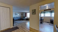 This single-floor residence offers a warm and inviting layout designed for comfortable everyday living. The living room serves as the heart of the home, featuring light-toned hardwood flooring that extends throughout the main living areas, lending a clean and cohesive feel to the space. Natural light filters in through multiple windows dressed with curtains, creating a bright and welcoming atmosphere. The living room provides ample space for seating and relaxation, with a textured ceiling adding subtle character overhead. A wide, open entryway connects the living room to the dining area, which is visible through a hallway passage. The dining space features its own window with decorative valance curtains, allowing additional natural light to flow in. Wall art and decorative accents are thoughtfully placed throughout, contributing to the space's lived-in charm. The layout promotes a smooth flow between rooms, making the home feel open and well-connected. Overall, the residence combines practical design with comfortable finishes, creating a functional and pleasant environment suitable for everyday living.
