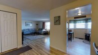 This single-floor residence offers a warm and inviting layout designed for comfortable everyday living. The living room serves as the heart of the home, featuring light-toned hardwood flooring that extends throughout the main living areas, lending a clean and cohesive feel to the space. Natural light filters in through multiple windows dressed with curtains, creating a bright and welcoming atmosphere. The living room provides ample space for seating and relaxation, with a textured ceiling adding subtle character overhead. A wide, open entryway connects the living room to the dining area, which is visible through a hallway passage. The dining space features its own window with decorative valance curtains, allowing additional natural light to flow in. Wall art and decorative accents are thoughtfully placed throughout, contributing to the space's lived-in charm. The layout promotes a smooth flow between rooms, making the home feel open and well-connected. Overall, the residence combines practical design with comfortable finishes, creating a functional and pleasant environment suitable for everyday living.