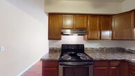 This single-floor residence offers a practical and cohesive layout designed for comfortable everyday living. The kitchen is a standout feature, showcasing rich, dark-toned wood cabinetry that extends to the upper walls, providing generous storage. Dark granite-style countertops complement the cabinetry, while a stainless steel electric range, stainless dishwasher, and a double-basin sink complete the functional workspace. Under-cabinet lighting adds a warm, practical touch to the cooking area. A decorative tile backsplash accent adds subtle character between the counters and upper cabinets. Warm hardwood flooring flows consistently throughout the space, lending a cohesive and inviting atmosphere. The living area is open and well-proportioned, bathed in natural light from large windows fitted with vertical blinds. A wall-mounted air conditioning unit and baseboard heating ensure year-round comfort. The bathroom features a full soaking tub with a shower, a wood-toned vanity with a white countertop, and a large mirror, all tied together with matching hardwood flooring. The overall layout, as revealed by the floorplan view, is efficiently organized, connecting the main living area, kitchen, and bathroom in a straightforward and accessible arrangement.