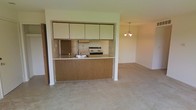 This single-floor space presents a practical and open layout that connects the kitchen and living areas seamlessly. The kitchen is defined by a wood-paneled peninsula counter that doubles as a breakfast bar, offering both workspace and a natural divider between the cooking and living zones. Upper cabinetry in a light cream finish provides ample storage, while a built-in range, overhead exhaust hood, refrigerator, and a stainless sink complete the functional setup. The kitchen's design emphasizes utility and accessibility, making it easy to navigate.

The adjoining living area is carpeted in a neutral, light tone that adds warmth and continuity throughout the space. An elegant chandelier with a classic multi-arm design serves as a focal point overhead, lending a touch of refinement to the otherwise understated room. The open sightlines between the kitchen and living area create a sense of spaciousness and encourage easy interaction across both zones. Neutral wall tones throughout keep the space feeling bright and versatile, providing a clean canvas well-suited for a variety of furnishing styles. Overall, the layout prioritizes functionality and ease of living within a cohesive, single-level floor plan.