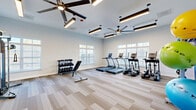 This fitness center offers a well-organized, single-level layout designed to support a wide range of workout routines. The space is finished with light-toned striped carpet flooring that runs throughout, contributing to a clean and contemporary aesthetic. Crisp white walls and generous natural light from multiple windows fitted with horizontal blinds create a bright, open atmosphere conducive to exercise.

The cardio zone is equipped with multiple treadmills and elliptical machines positioned along the windowed wall, allowing users to work out with outdoor views. A dedicated free weights area features a multi-tier dumbbell rack paired with an adjustable weight bench, providing options for strength training. A multi-functional cable machine station supports a broad spectrum of resistance exercises and is situated toward one end of the room.

A separate functional training area offers open floor space alongside a storage rack stocked with stability balls, medicine balls, and resistance bands, making it suitable for stretching, balance work, or group exercise. Overhead, large ceiling fans with integrated linear pendant lighting ensure consistent air circulation and illumination throughout. The overall design is modern and purposeful, balancing equipment density with sufficient open space for comfortable movement.