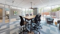 This contemporary single-floor office space is thoughtfully designed to balance collaboration and focused work. The layout features a series of glass-partitioned areas framed in sleek dark metal, creating an open yet structured feel that allows natural light to flow throughout the interior. Floor-to-ceiling windows look out onto an adjacent outdoor patio area furnished with seating, seamlessly connecting the indoors with the exterior environment.

The workspace includes a dedicated collaborative meeting area centered around a solid wood high-top table paired with ergonomic task chairs, ideal for team discussions or brainstorming sessions. A pendant light fixture anchors the meeting space, while abstract artwork on the walls adds a refined, creative touch. Individual workstations equipped with mesh-back ergonomic chairs are positioned along the perimeter, offering focused work zones within the open plan.

A comfortable lounge area features a cushioned sofa set against a textured gray accent wall adorned with bold abstract paintings and a wall-mounted flat-screen display, providing a space for informal gatherings or presentations. Throughout the space, a patterned modular carpet in cool blue and gray tones unifies the design, complementing the neutral palette of the walls and furnishings. The overall ambiance is professional yet inviting, reflecting a modern approach to workplace design.