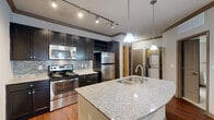 This 1-bedroom, 1-bathroom residence is thoughtfully designed with an open-concept layout that seamlessly connects the kitchen, dining, and living areas. The kitchen is a standout feature, showcasing dark espresso cabinetry, a mosaic tile backsplash, stainless steel appliances including a refrigerator, range, and built-in microwave, as well as a built-in wine rack. A curved granite-topped island with a stainless steel sink and pendant lighting serves as both a functional workspace and a natural focal point of the space. Rich hardwood-style flooring flows throughout the main living areas, complemented by crown molding and ceiling fans. The spacious living room is bright and airy, featuring French doors that invite natural light and provide access to an outdoor area. The bedroom is carpeted for comfort, offering a ceiling fan, natural light from windows with blinds, and a walk-in closet. The bathroom features a full tub-shower combo with floor-to-ceiling tile surround, granite vanity countertop, and coordinating dark cabinetry. A stacked in-unit washer and dryer is conveniently located within the residence, adding to its practical appeal. Crown molding and track lighting throughout lend a polished, cohesive finish to the entire space.