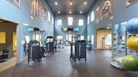 This fitness center offers an expansive, single-floor layout designed to accommodate a full range of workout routines. The space is immediately distinguished by its soaring double-height ceilings, painted a deep slate blue that contrasts with the lighter blue walls, creating a sleek and energizing atmosphere. Recessed lighting and a decorative pendant fixture illuminate the room from above, while rows of arched windows along both the upper and lower portions of the walls flood the space with abundant natural light.

The facility is outfitted with a comprehensive selection of Magnum strength-training machines, strategically arranged to allow comfortable movement throughout the floor. Cardio equipment lines the background near the entrance doors, offering variety for users. Large-format athletic photography adorns the walls, adding a motivational aesthetic to the environment. Multiple flat-screen televisions are mounted near the front, ensuring entertainment options during workouts.

Full-length mirrors along one wall enhance the sense of openness while serving a functional purpose. The durable flooring, featuring interlocking rubber mat sections, provides safety and comfort underfoot. Overall, the space balances functionality with an inviting, professional design.