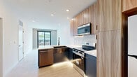 Located at 1923 Broadway Blvd in Kansas City, Missouri, this thoughtfully designed one-bedroom, one-bathroom unit offers a contemporary urban living experience. The open-concept layout seamlessly connects the kitchen and living area, finished with light hardwood-style flooring that extends throughout. The kitchen is equipped with stainless steel appliances, including a gas range, microwave, and refrigerator, complemented by warm wood cabinetry and a marble-style backsplash that adds a refined touch. A bold charcoal entry door sets a modern tone upon arrival. The living area is bathed in natural light through large floor-to-ceiling sliding glass doors that open to a private balcony, offering sweeping views of the Kansas City skyline and surrounding urban streetscape. The bedroom features soft carpeting, a ceiling fan, and a double-door closet, providing both comfort and storage. A second door leads conveniently to the bathroom, which showcases striking floor-to-ceiling navy vertical tile in a glass-enclosed walk-in shower, paired with elegant marble-look tile flooring and matte black fixtures throughout. Clean white walls, recessed lighting, and modern hardware complete the refined, contemporary aesthetic of this well-appointed urban residence.