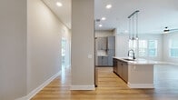 This single-floor residence showcases a thoughtfully designed open-concept layout that seamlessly connects the kitchen, dining, and living areas. The kitchen is a standout feature, centered around a white quartz-topped island equipped with a matte black sink and faucet. Gray shaker-style cabinetry lines the walls, paired with a subway tile backsplash and stainless steel appliances, including a dishwasher and refrigerator. A trio of globe pendant lights suspended from a black track adds a contemporary finishing touch above the island. Recessed lighting and large windows allow generous natural light to fill the space, with views of surrounding greenery visible from the living area. Light hardwood flooring flows consistently throughout, lending warmth and visual continuity across all rooms. The bedrooms are well-proportioned, featuring neutral wall tones, ceiling fans, and ample natural light. One bedroom offers a walk-in closet with wire shelving for organized storage. The bathroom is finished with a gray vanity, white quartz countertop, an illuminated mirror, matte black fixtures, and a full tub-shower combination enclosed by marble-look tile surround.