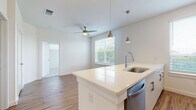 This single-floor residence offers a thoughtfully designed open-concept layout with clean, modern finishes throughout. The kitchen serves as a central highlight, featuring crisp white cabinetry, a large quartz island with an undermount stainless steel sink, and a full suite of stainless steel appliances including a range, microwave, refrigerator, and dishwasher. Stylish pendant lighting hangs above the island, complementing the bright, airy atmosphere created by multiple windows dressed with horizontal blinds. Warm wood-look flooring flows through the kitchen and living areas, adding a natural contrast to the white palette. The living area extends naturally from the kitchen and includes a ceiling fan, making it a comfortable and functional shared space. The bedroom is carpeted for added comfort and benefits from a large window that invites ample natural light and outdoor views. The bathroom is finished to a high standard, featuring a wide quartz vanity with white cabinetry, a large frameless mirror, and a full tub and shower surround clad in dramatic marble-look tile with bold veining. Recessed lighting and wood-look flooring complete the bathroom's refined aesthetic. Overall, this residence combines modern design with practical functionality.