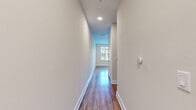 This single-floor space offers a cohesive, modern design highlighted by warm hardwood flooring that runs throughout. A long, well-lit hallway with recessed lighting and clean white walls serves as the central corridor connecting the various areas of the unit, with a smart thermostat mounted along its length.

The kitchen is a standout feature, equipped with full stainless steel appliances including a refrigerator, electric range, over-the-range microwave, and dishwasher. White shaker-style cabinetry extends to the ceiling, providing generous storage, while a classic subway tile backsplash and under-cabinet lighting add refined detail.

The open living area is defined by neutral tones and ample wall space, with natural light filtering in through large windows fitted with horizontal blinds. The bedroom similarly benefits from a wide window offering outdoor views of greenery, creating a bright and comfortable atmosphere.

The bathroom features a tub-shower combination with large-format white tile surround, complemented by matte black fixtures that lend a contemporary contrast. Barn-style sliding doors, visible in the floor plan, add a distinctive architectural touch throughout the space.