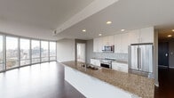 This single-floor unit showcases a thoughtfully designed open layout with a strong emphasis on natural light and urban views. The living area is a standout feature, boasting a dramatic curved wall of floor-to-ceiling windows that frame expansive city skyline views and flood the space with daylight. Rich, dark hardwood flooring anchors the open-concept living and dining areas, lending warmth to the contemporary aesthetic.

The kitchen is well-appointed with crisp white cabinetry extending to the ceiling, a light glass tile backsplash, and speckled granite countertops. A generously sized island with a double undermount sink serves as both a functional workspace and a natural gathering point. Stainless steel appliances, including a range, built-in microwave, and refrigerator, complete the modern culinary setup.

The bedrooms feature neutral carpeting and large windows with city views, offering a quiet retreat from the open living spaces. One bedroom benefits from an impressive floor-to-ceiling sliding glass window wall with access to an outdoor area.

The bathrooms are finished consistently with granite-topped white vanities, large mirrors, soaker tub and shower combinations, and light tile flooring, maintaining the unit's clean, modern character throughout.