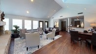 This single-story clubhouse offers an inviting and well-appointed gathering space designed for comfort and community use. The main living area immediately draws attention with its dramatic vaulted ceiling, which creates an expansive, airy atmosphere throughout the room. A floor-to-ceiling stacked stone fireplace with a bold wooden mantel serves as the focal centerpiece, adding warmth and rustic elegance to the space. Rich, dark hardwood flooring runs throughout the main area, complementing the neutral gray walls and creating a cohesive, sophisticated aesthetic.

The seating area is generously sized, accommodating multiple conversation groupings with tufted armchairs and leather sofas arranged atop a patterned area rug. Large windows with white blinds line the walls, allowing abundant natural light to fill the room while offering views of the surrounding outdoors. A bar-height counter and seating area connect to an adjacent kitchen, which is equipped with ample cabinetry, countertop space, a full-size refrigerator, microwave, and sink — providing practical amenities for events and gatherings. The overall ambiance is warm, comfortable, and well-suited for hosting community activities.