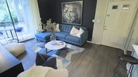 This single-floor space showcases a thoughtfully designed living area that blends modern aesthetics with a cozy, inviting atmosphere. The room is anchored by rich, dark hardwood-style flooring that runs throughout, providing a warm and grounded foundation for the space. A striking deep charcoal accent wall serves as a dramatic backdrop, creating depth and contrast that defines the room's contemporary character.

The living area is centered around a tufted blue sectional sofa, offering both comfort and visual appeal. A round white coffee table sits atop a geometric patterned area rug in muted tones of blue, grey, and white, adding texture and dimension to the floor space. A glass vase with dried branches provides an organic, decorative touch to the coffee table arrangement.

Wall décor includes a large framed floral wreath artwork that complements the dark accent wall beautifully. Natural light filters into the space through what appears to be a glass door or large window along the perimeter, brightening the room and enhancing its open feel. The overall ambiance is sophisticated, modern, and carefully curated, making it both functional and visually compelling.