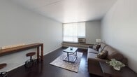 Located at 100 Fairview Square in Ithaca, NY, this single-floor residence offers a thoughtfully organized layout with distinct living, sleeping, kitchen, and bathroom spaces. The living room is a welcoming area highlighted by large windows with vertical blinds that allow generous natural light to fill the room. Dark hardwood-style flooring runs throughout, adding warmth and contrast against the light gray walls. A window-unit air conditioner and baseboard heating ensure year-round comfort. The kitchen is efficiently designed with rich dark cabinetry, marble-style countertops, stainless steel appliances including a gas range and built-in microwave, and a functional sink — all contributing to a modern, practical cooking space. A raised countertop peninsula separates the kitchen from the adjacent dining area. The bedroom is a quiet, simple retreat with the same dark flooring, soft gray walls, a window with vertical blinds, and baseboard heating. The full bathroom features white subway tile walls, a pedestal sink with a modern faucet, a mirror medicine cabinet, a chrome towel rack, and a full bathtub with a shower curtain, creating a clean and functional space. Overall, the residence presents a clean, modern aesthetic with practical amenities throughout.