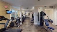 This fitness center offers a thoughtfully designed workout space spread across a single level, divided into distinct zones that accommodate a full range of exercise activities. The layout is organized to separate cardio equipment from strength training areas, allowing for efficient use of the space without congestion.

The cardio zone is anchored by multiple treadmills, elliptical trainers, and a stair climber, all positioned near large bay windows that allow generous natural light to filter in and provide outdoor views during workouts. Wall-mounted flat-screen televisions are positioned for easy viewing from the cardio machines, enhancing the workout experience.

The strength training area features a multi-station cable machine, leg press, and weight bench, along with stability balls and free weights. Structural columns with classic detailing define the transition between zones, contributing to the architectural character of the space.

Throughout, warm hardwood-style flooring, crisp white crown molding, and neutral walls create a clean, polished aesthetic. Ceiling fans in both zones promote air circulation. Framed vintage athletic and Olympic-themed posters serve as decorative accents, reinforcing the fitness-focused atmosphere. The overall ambiance is bright, functional, and well-maintained.