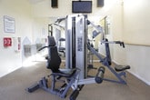 This fitness center is a thoughtfully designed exercise facility offering a comprehensive range of workout equipment across a single-level, open layout. The space is outfitted with multiple Precor cardio machines, including treadmills, an elliptical, and a rowing machine, alongside a multi-station cable strength training system that supports a full-body workout routine. The flooring consists of neutral, wall-to-wall carpet, providing a comfortable underfoot surface throughout the space. Warm yellow-toned walls and recessed lighting create a bright and energizing atmosphere, complemented by ceiling fans that promote air circulation. A wall-mounted flat-screen television and mounted speakers add an entertainment element to support engaging workout sessions. Large black-framed windows along one wall allow abundant natural light to filter in and offer views of an adjacent outdoor patio area. Full-length mirrors along one wall visually expand the space and allow users to monitor their form during exercise. The facility also features a raised secondary area accessible by steps, visible through an interior window, which adds a sense of architectural depth. A clearly posted exercise room rules sign reflects an organized, community-oriented environment. Overall, the space is clean, functional, and well-maintained, catering to a wide range of fitness needs.