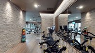 This contemporary fitness center combines functionality with refined design, creating an inviting environment for a comprehensive workout experience. The space features striking exposed brick walls that lend an industrial-chic aesthetic, complemented by warm hardwood flooring that runs throughout. Recessed and ambient lighting illuminates the facility, while ceiling fans provide additional airflow and comfort during intense training sessions.

The cardio zone is well-appointed with a row of stationary cycling bikes, including LeMond and Expresso interactive models equipped with digital screens for guided and performance-tracked rides. Full-length mirrors line the walls, enhancing the sense of space and allowing users to monitor their form during workouts.

The strength training area is visible beyond, offering a variety of resistance machines and benches suitable for a full-body training regimen. A dedicated medicine ball rack, stocked with a colorful assortment of weighted balls, supports functional fitness and cross-training routines. Structural columns with warm-toned brick cladding punctuate the floor plan, adding architectural character throughout.

Overall, the fitness center delivers a polished and professional atmosphere that balances modern equipment with thoughtful interior design, making it suitable for users of all fitness levels.