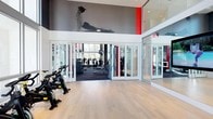 This fitness facility is designed with both functionality and aesthetics in mind, offering a well-organized layout that caters to a variety of workout styles. The space is divided into distinct zones, separated by expansive glass-paneled folding doors that allow for flexible configuration while maintaining an open, airy feel throughout.

The cycling area features light hardwood-style flooring and is positioned near large windows that invite natural light, creating an energizing environment for cardio training. A striking large-scale black-and-white mural of a figure in a yoga pose adorns the upper wall, adding a dynamic visual element that reflects the facility's fitness-focused identity. Bold red accents throughout the space complement the modern, high-contrast color palette of white and grey.

Beyond the glass dividers lies the main strength and cardio training area, furnished with an array of weight machines and equipment arranged across carpeted flooring. The soaring ceiling height enhances the sense of spaciousness, while large windows along the perimeter allow for views of the surrounding outdoor environment. The overall ambiance is clean, modern, and motivating, suited for both individual and group exercise routines.