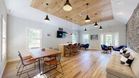 Located at 9 Covered Bridge Rd in Newtown, CT, this single-floor space offers a well-appointed layout comprising a combined living and dining room alongside a fully equipped kitchen. The most striking architectural feature is a floor-to-ceiling stacked stone accent wall in the living area, rendered in layered tones of grey, taupe, and rust that lend the space a rustic yet contemporary character. Warm-toned hardwood flooring runs throughout, complementing the neutral grey walls and white trim. The living area is open and inviting, with recessed lighting and ample room for comfortable seating. The dining section is bathed in natural light from large double-hung windows that frame views of the surrounding greenery, creating a bright and airy atmosphere. A wood-paneled ceiling detail adds warmth and texture to the dining and lounge zone, while pendant lighting provides focused illumination. The kitchen features crisp white cabinetry, granite countertops, a white tile backsplash, stainless steel appliances, and open wood shelving. A kitchen island with bar seating in leather-toned chairs enhances the space's functionality. French doors at one end of the living area connect the interior to the outdoors, further enhancing the sense of openness throughout.