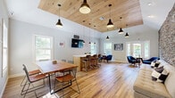 Located at 9 Covered Bridge Rd in Newtown, CT, this single-floor space offers a well-appointed layout comprising a combined living and dining room alongside a fully equipped kitchen. The most striking architectural feature is a floor-to-ceiling stacked stone accent wall in the living area, rendered in layered tones of grey, taupe, and rust that lend the space a rustic yet contemporary character. Warm-toned hardwood flooring runs throughout, complementing the neutral grey walls and white trim. The living area is open and inviting, with recessed lighting and ample room for comfortable seating. The dining section is bathed in natural light from large double-hung windows that frame views of the surrounding greenery, creating a bright and airy atmosphere. A wood-paneled ceiling detail adds warmth and texture to the dining and lounge zone, while pendant lighting provides focused illumination. The kitchen features crisp white cabinetry, granite countertops, a white tile backsplash, stainless steel appliances, and open wood shelving. A kitchen island with bar seating in leather-toned chairs enhances the space's functionality. French doors at one end of the living area connect the interior to the outdoors, further enhancing the sense of openness throughout.