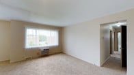 This 2-bedroom, 1-bathroom single-floor unit offers a clean, functional layout designed for comfortable everyday living. The living area serves as the central gathering space, characterized by neutral beige walls and wall-to-wall carpeting that creates a warm, understated aesthetic. A large window fitted with vertical blinds allows generous natural light to fill the room while framing views of the surrounding greenery and adjacent buildings. A wall-mounted air conditioning unit sits beneath the window, providing dedicated climate control directly within the living space. The smooth white ceiling and simple, unadorned walls give the room a bright and open feel, making it easy to personalize with furnishings and décor. A wide entryway or hallway passage is visible along one wall, indicating a straightforward, accessible flow between the living area and the rest of the unit. The overall ambiance is clean and move-in ready, with a practical design that prioritizes usability. The neutral palette and carpeted floors throughout contribute to a cohesive, quiet atmosphere suited for a range of lifestyles.