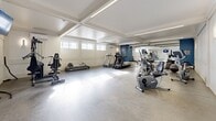 Located at 4555 Av Bonavista in Montréal, this fitness facility offers a thoughtfully designed exercise space spread across a single floor. The layout is accessed through a clean, well-lit hallway finished in crisp white with wainscoting panelling and recessed ceiling lighting, creating a welcoming and organized entry to the space.

The exercise room is generously sized, featuring a bright, open layout with a coffered white ceiling and integrated linear lighting that evenly illuminates the entire area. A wall-mounted air conditioning unit ensures year-round comfort. Natural light filters in through a row of horizontal windows positioned high along one wall, complementing the interior lighting. The floor is finished with smooth, light-toned hardwood-style flooring throughout.

Equipment is well distributed across the room and includes treadmills, an elliptical trainer, a recumbent stationary bike, a cable machine, a weight bench, and a dumbbell rack with a broad wall-mounted mirror. A ceiling-mounted television is installed in one section of the room, offering entertainment during workouts. A bold blue accent wall with an arched detail adds a distinctive visual element. Informational signage, a wall clock, and sanitation supplies are also present, reflecting attention to functionality and hygiene. The overall ambiance is clean, modern, and professional.