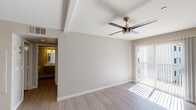 This single-floor residence offers a clean, modern aesthetic throughout, with light-toned luxury vinyl plank flooring and neutral warm-gray walls creating a cohesive and inviting atmosphere. The living area is a standout space, featuring a tray ceiling detail, a ceiling fan with integrated lighting, and a sliding glass door fitted with vertical blinds that floods the room with natural light and provides access to an outdoor balcony. The galley-style kitchen is thoughtfully designed with white shaker cabinetry, gray quartz-style countertops, and a white subway tile backsplash. Stainless steel appliances — including a gas range, microwave, and refrigerator — are complemented by track lighting overhead and a single-basin sink, making the kitchen both functional and visually appealing. The bedroom is generously proportioned, featuring its own ceiling fan, a window with horizontal blinds for natural light and privacy, and easy access to the bathroom. The bathroom is finished with a full tub and shower combination framed in white tile, brushed nickel fixtures, and wood-look flooring that ties into the overall design language of the home. The layout, as visible from the overhead dollhouse view, flows efficiently between spaces, offering a practical and comfortable living environment.