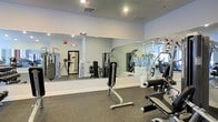 This single-level fitness center is designed to provide a functional and comfortable workout environment. The space is outfitted with a variety of strength training machines and equipment, including cable machines and adjustable benches, catering to a wide range of exercise needs. A large floor-to-ceiling mirror spans one wall, visually expanding the space while allowing users to monitor their form during workouts. The flooring consists of dark rubber matting, which provides durability, impact absorption, and traction underfoot. The walls are finished in a clean, neutral light gray, contributing to a bright and focused atmosphere. Recessed ceiling lighting ensures even, well-distributed illumination throughout the room. A window on one side allows natural light to enter, adding to the open feel of the space. The layout is thoughtfully organized to allow adequate circulation between equipment. A fire alarm and ventilation systems are visibly integrated into the ceiling and walls, meeting safety and comfort standards. Overall, the fitness center offers a clean, practical, and well-maintained environment suited for regular exercise.